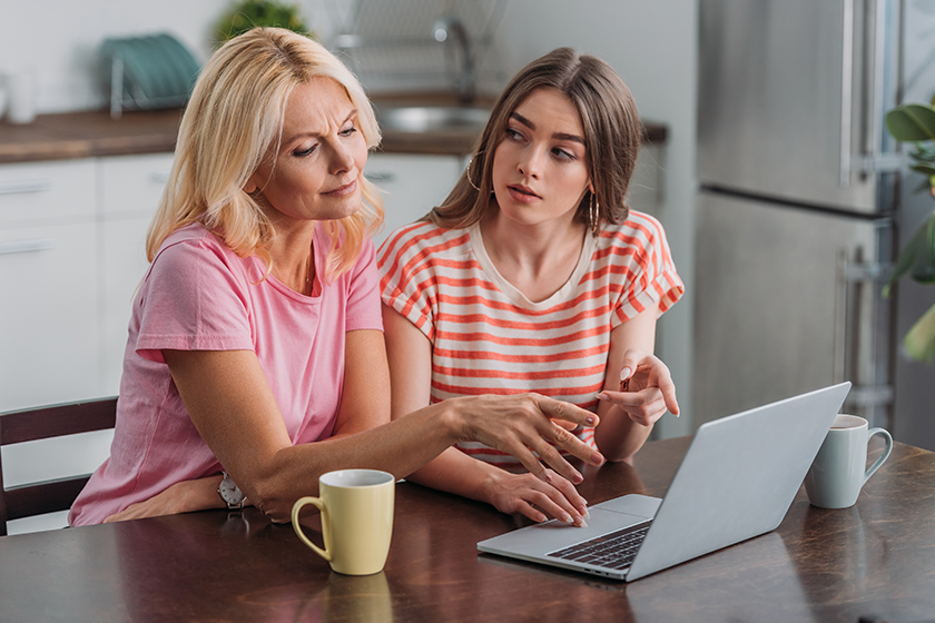 Mother and daughter pointing with fingers at laptop while sitting at kitchen table 