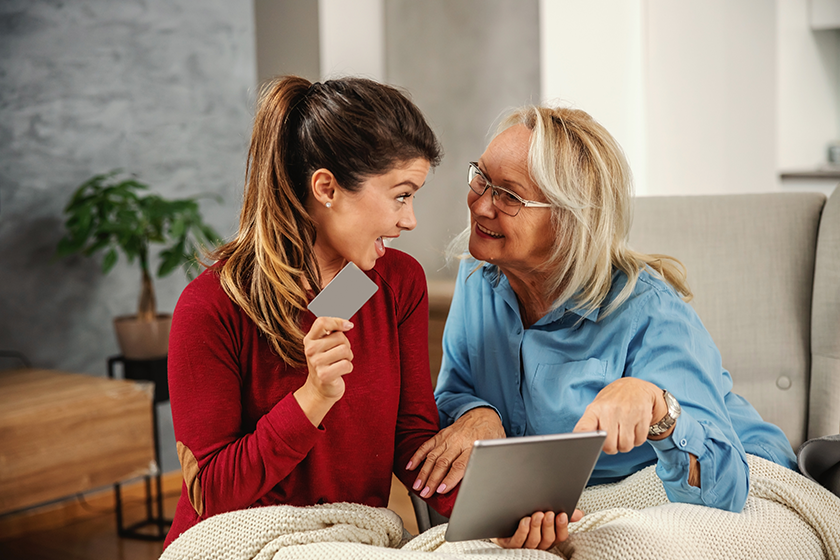 Smiling blond senior woman sitting ina chair and looking at tablet. Her daughter sitting next to her, holding tablet and credit card. They are about to shop online. 